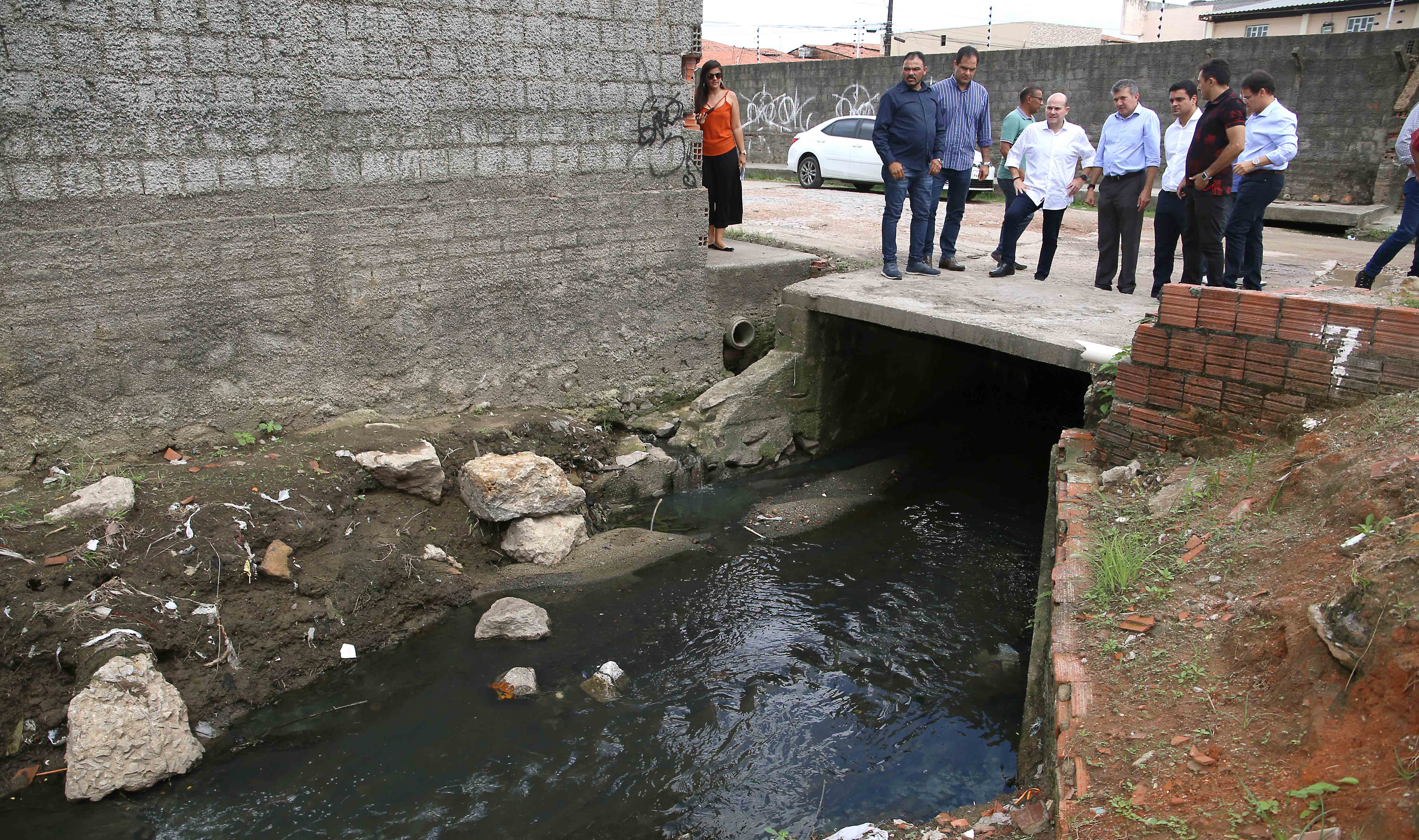 prefeito e pessoas em pé em cima de canal com água suja passando embaixo